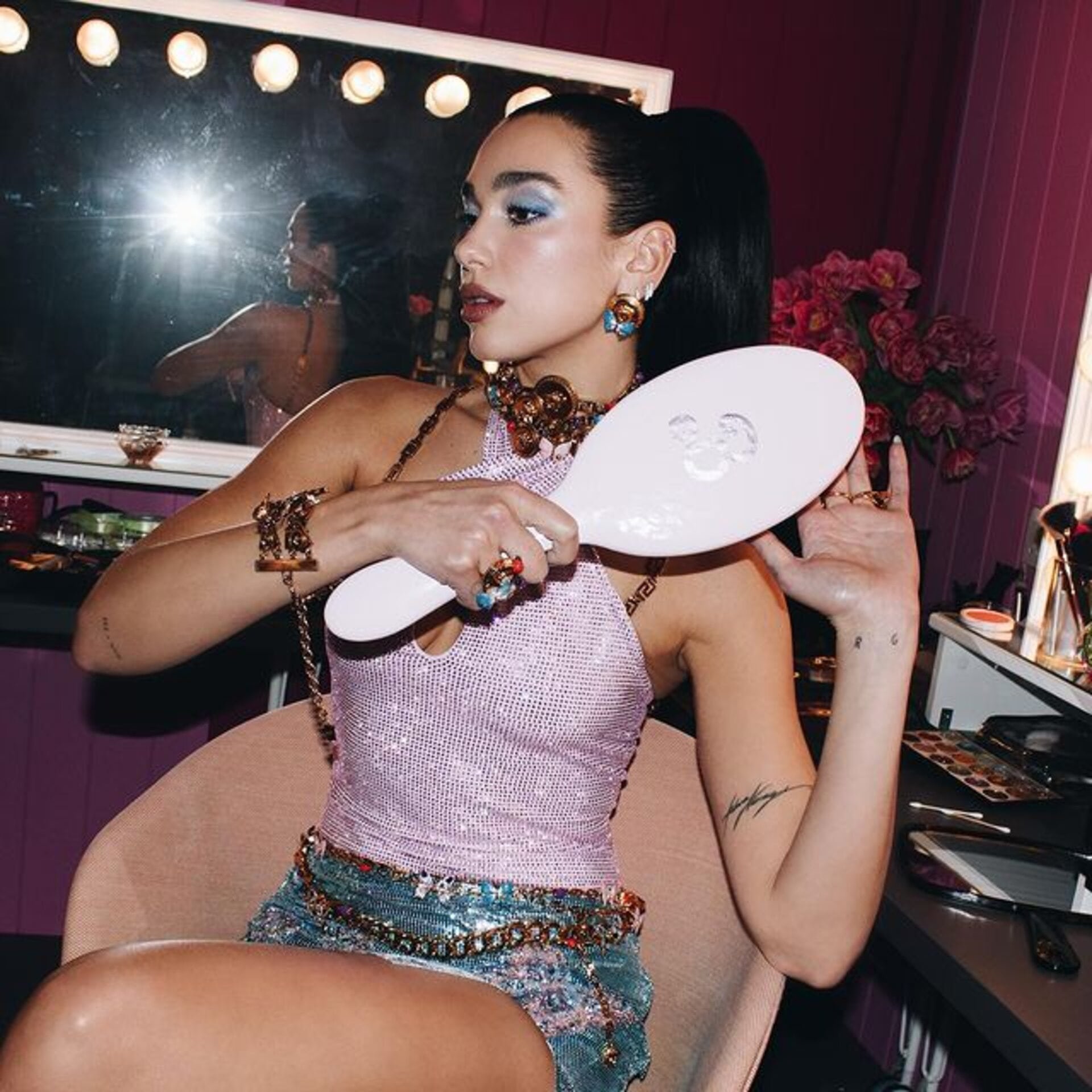 Adult woman brushing her hair in front of a mirror surrounded by light bulbs, wearing a pink top and jeweled belt and skirt.