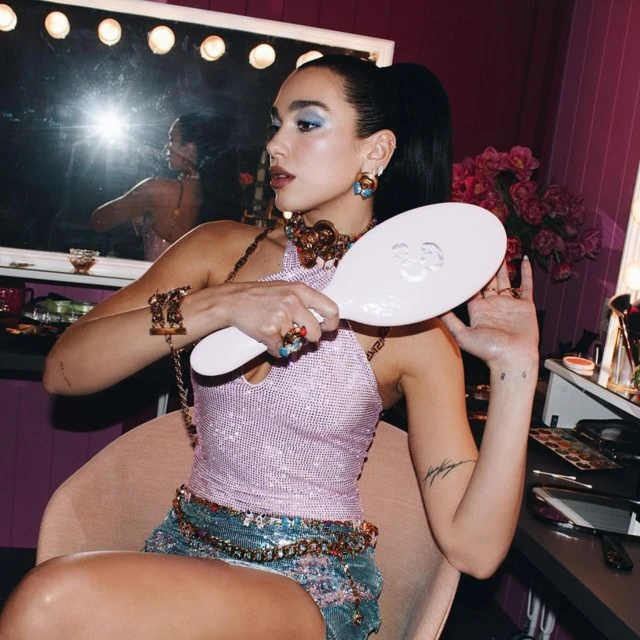 Adult woman brushing her hair in front of a mirror surrounded by light bulbs, wearing a pink top and jeweled belt and skirt.