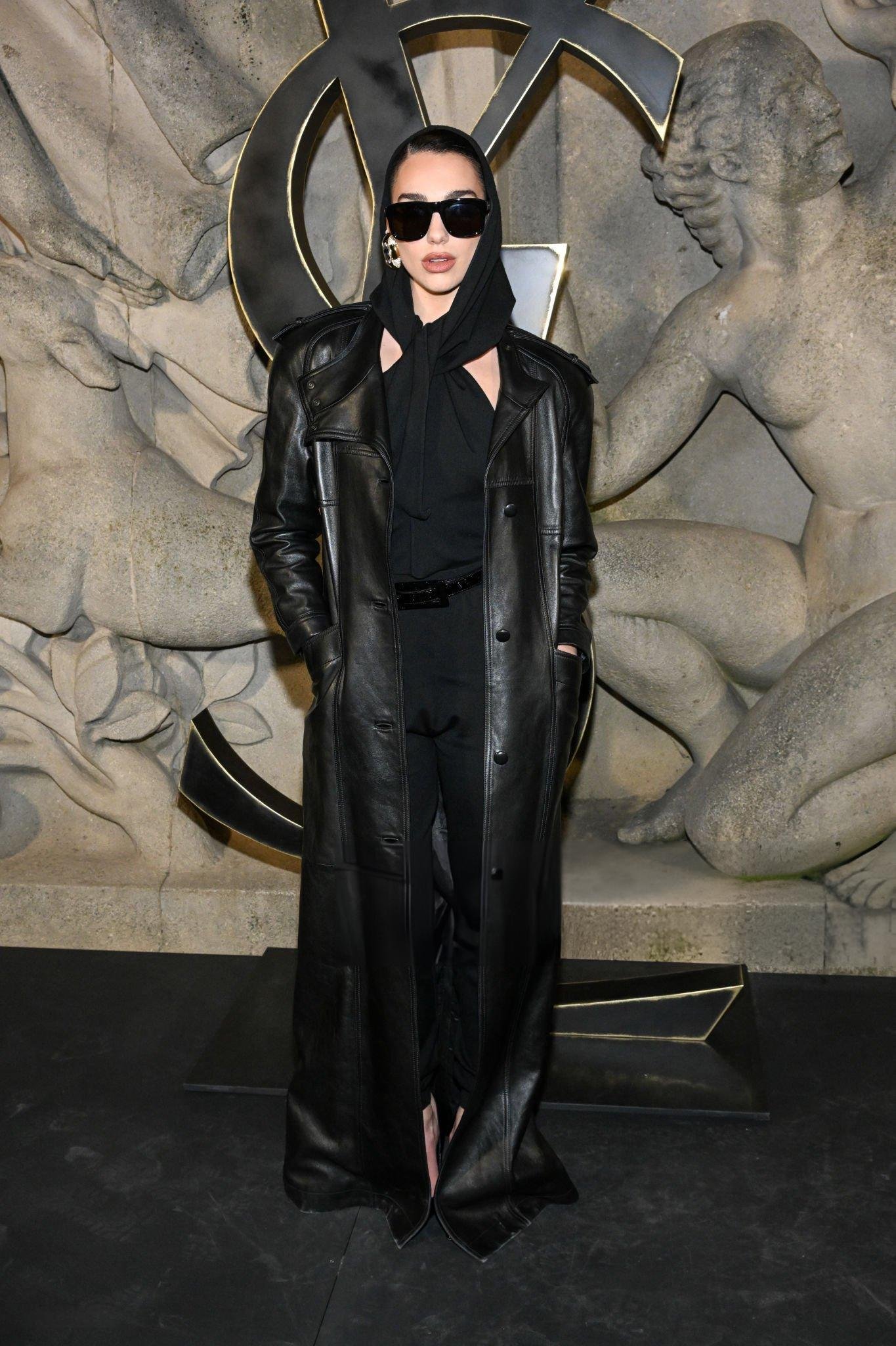 Woman in black leather trench coat and sunglasses poses in front of carved stone background featuring YSL logo and human figures.