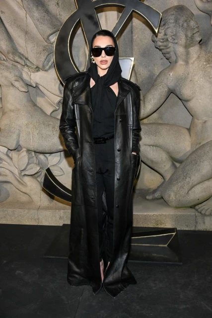 Woman in black leather trench coat and sunglasses poses in front of carved stone background featuring YSL logo and human figures.