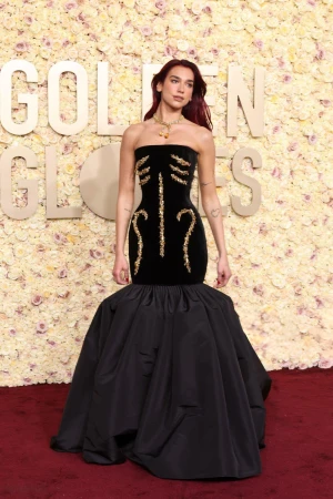 Dua Lipa poses on a red carpet wearing a black strapless gown with gold embellishments and a voluminous skirt in a floral setting.