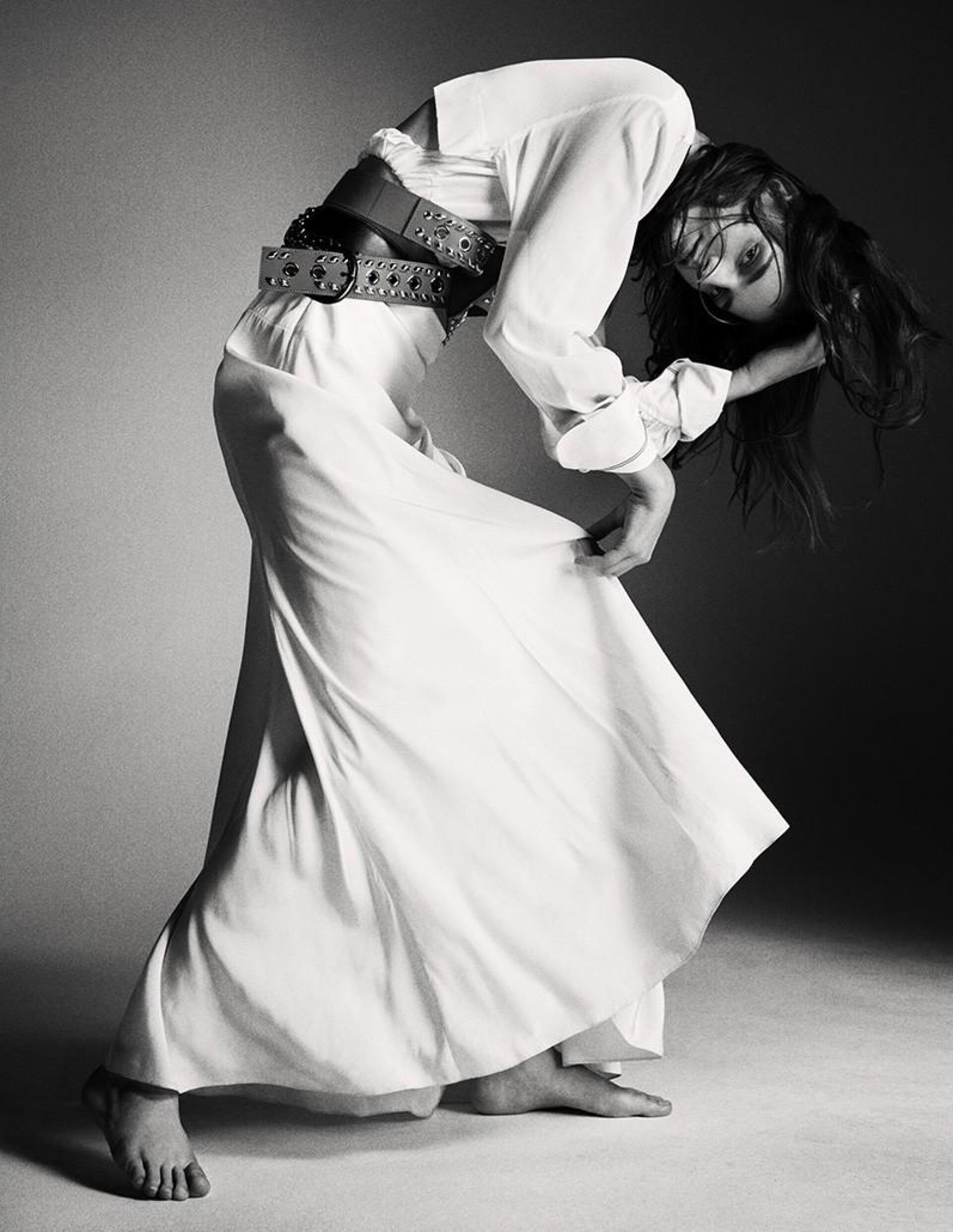 Black and white fashion shot of a woman in a white dress and wide belt, bending backward in a studio with soft, diffused lighting.