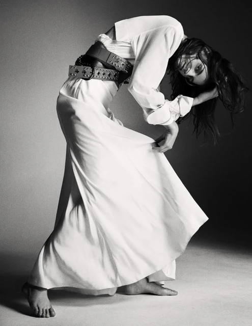 Black and white fashion shot of a woman in a white dress and wide belt, bending backward in a studio with soft, diffused lighting.