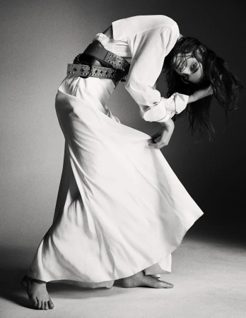 Black and white fashion shot of a woman in a white dress and wide belt, bending backward in a studio with soft, diffused lighting.