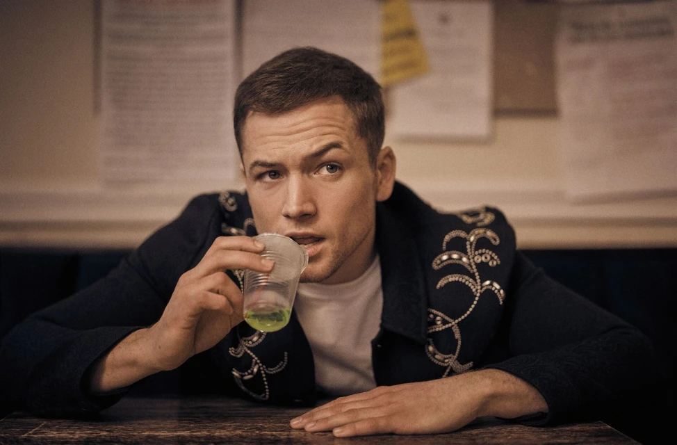 Close-up of a man in a dark jacket with floral embellishments drinking a green liquid from a plastic cup while seated at a wooden table.