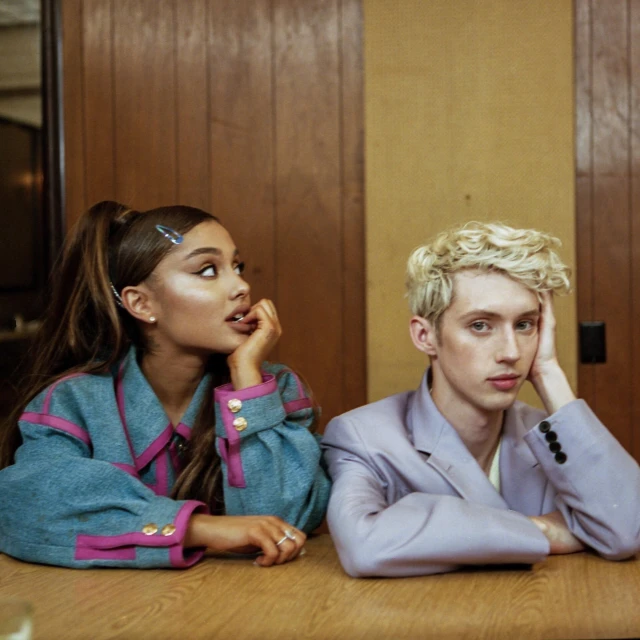 Candid shot of Ariana Grande and Troye Sivan seated at a table, wearing stylish outfits, and looking off-camera in an indoor setting.
