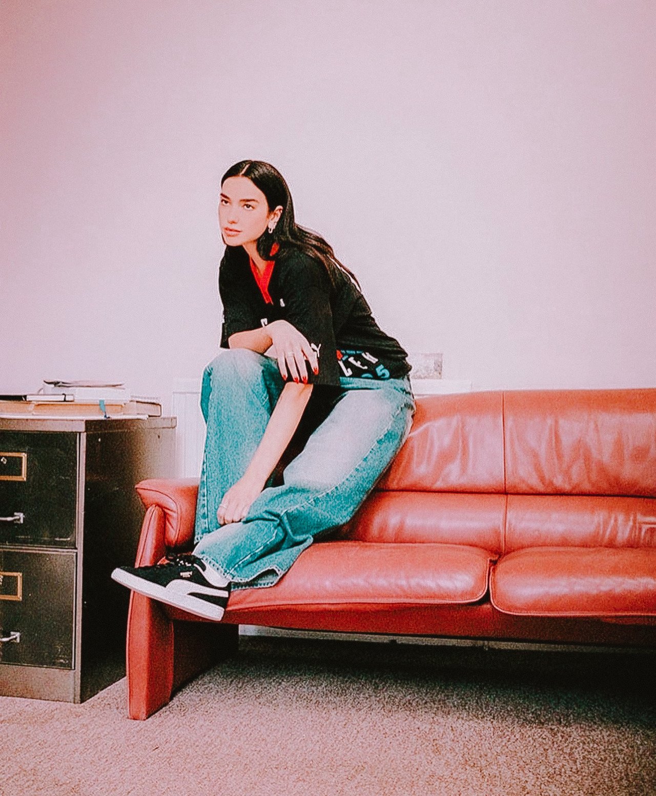 Brunette woman in jeans and Puma sneakers sits on a vintage red couch in a room with neutral carpet and a file cabinet.