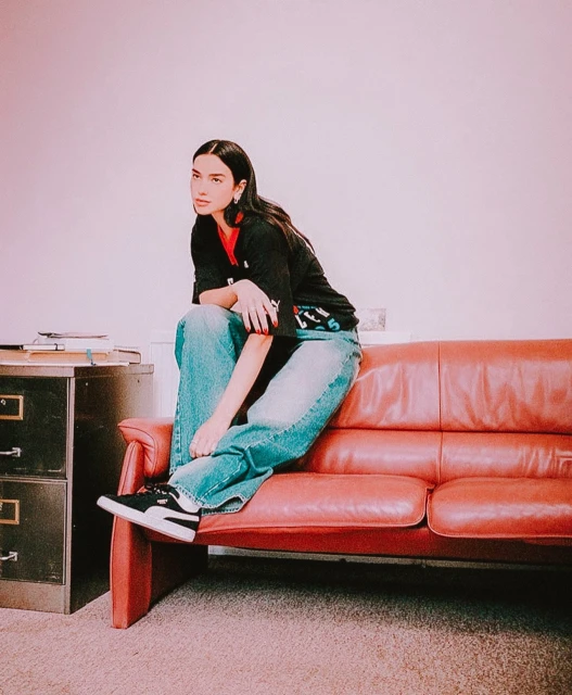 Brunette woman in jeans and Puma sneakers sits on a vintage red couch in a room with neutral carpet and a file cabinet.
