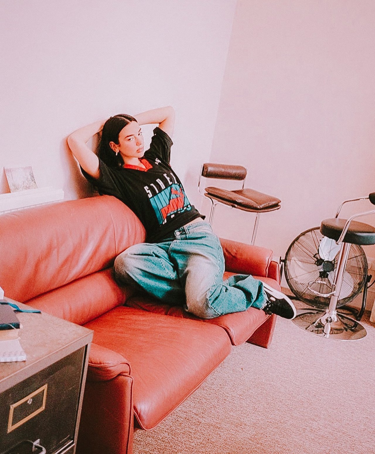 Young adult woman with dark hair lounges on a red couch in casual denim, black t-shirt indoors, with a vintage aesthetic.