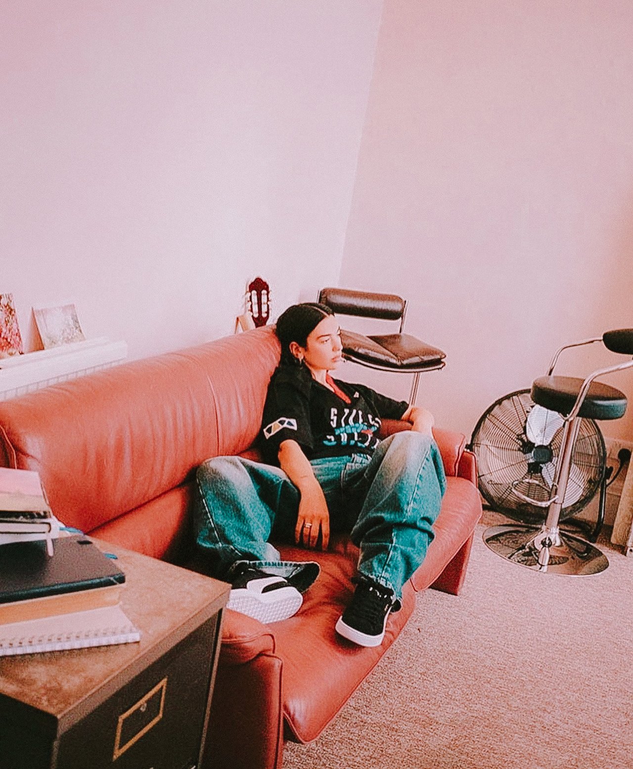 Indoor portrait of a relaxed woman in a black shirt and baggy jeans lounging on a red couch. Retro decor and soft pink lighting.
