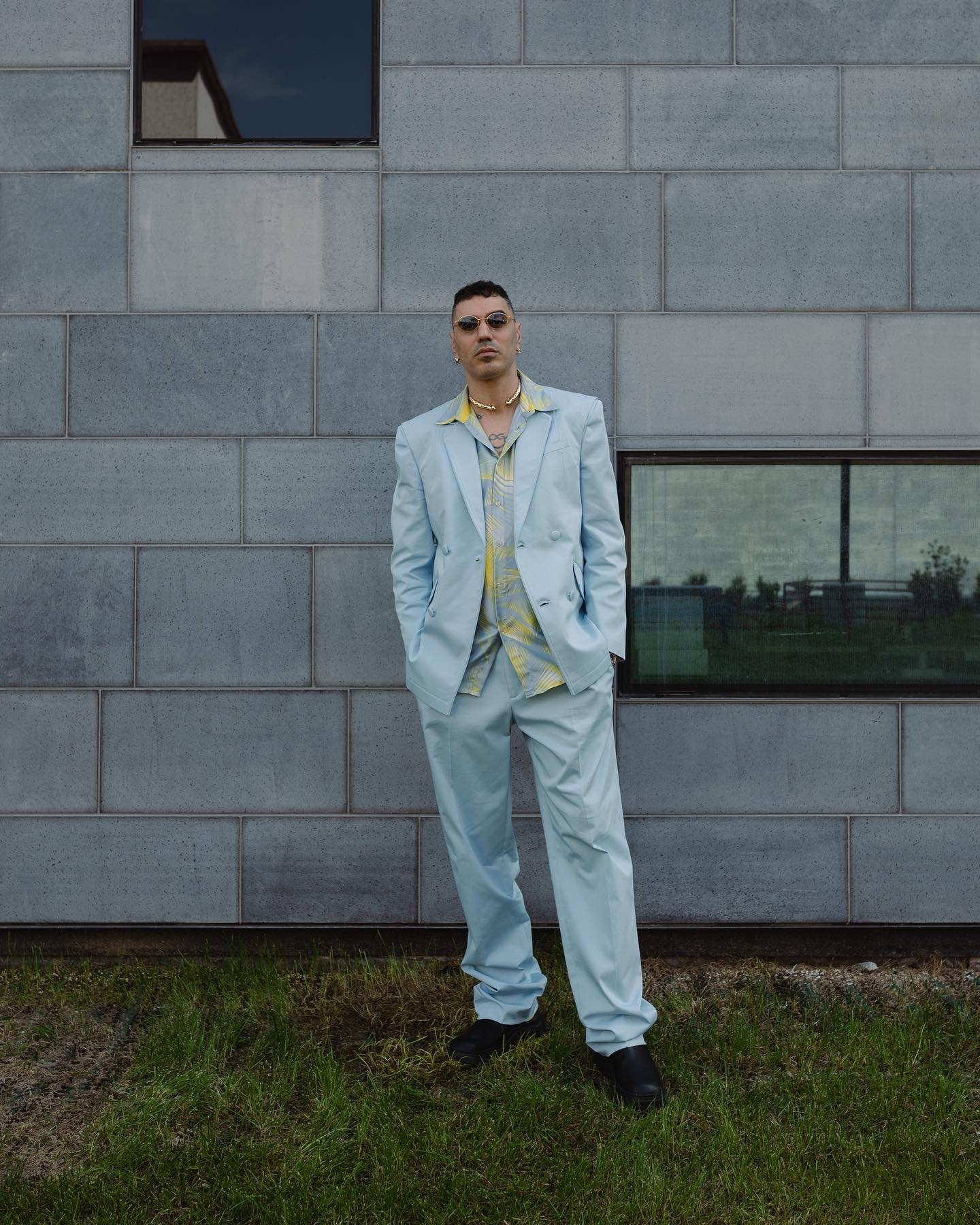 Man wearing a light blue suit and patterned shirt standing against a grey tiled building exterior