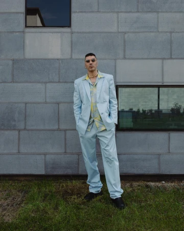 Man wearing a light blue suit and patterned shirt standing against a grey tiled building exterior