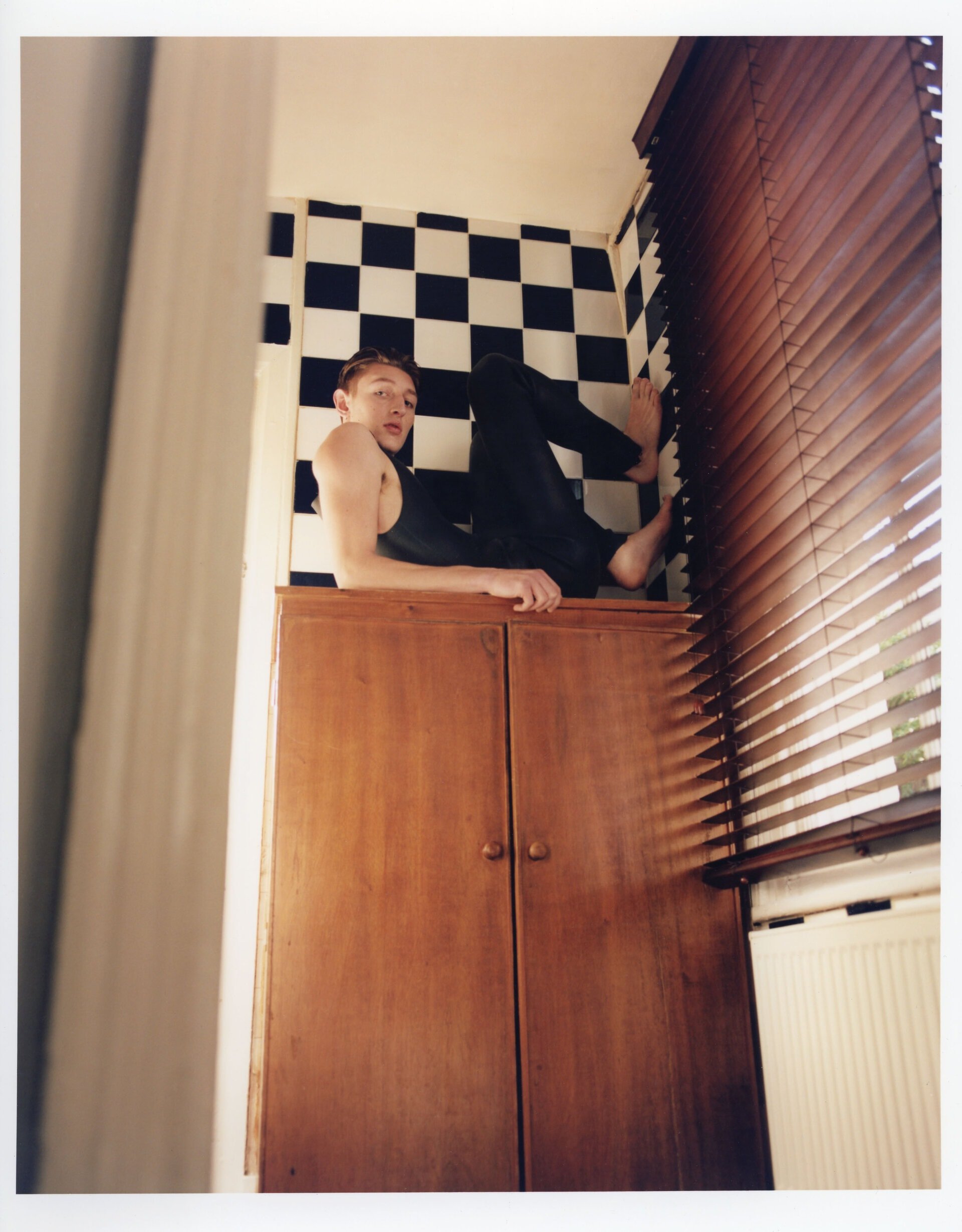 Adult male casually reclines atop a wooden cabinet indoors, featuring black and white checkered tile backsplash and slatted window blinds.