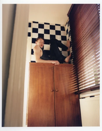 Adult male casually reclines atop a wooden cabinet indoors, featuring black and white checkered tile backsplash and slatted window blinds.