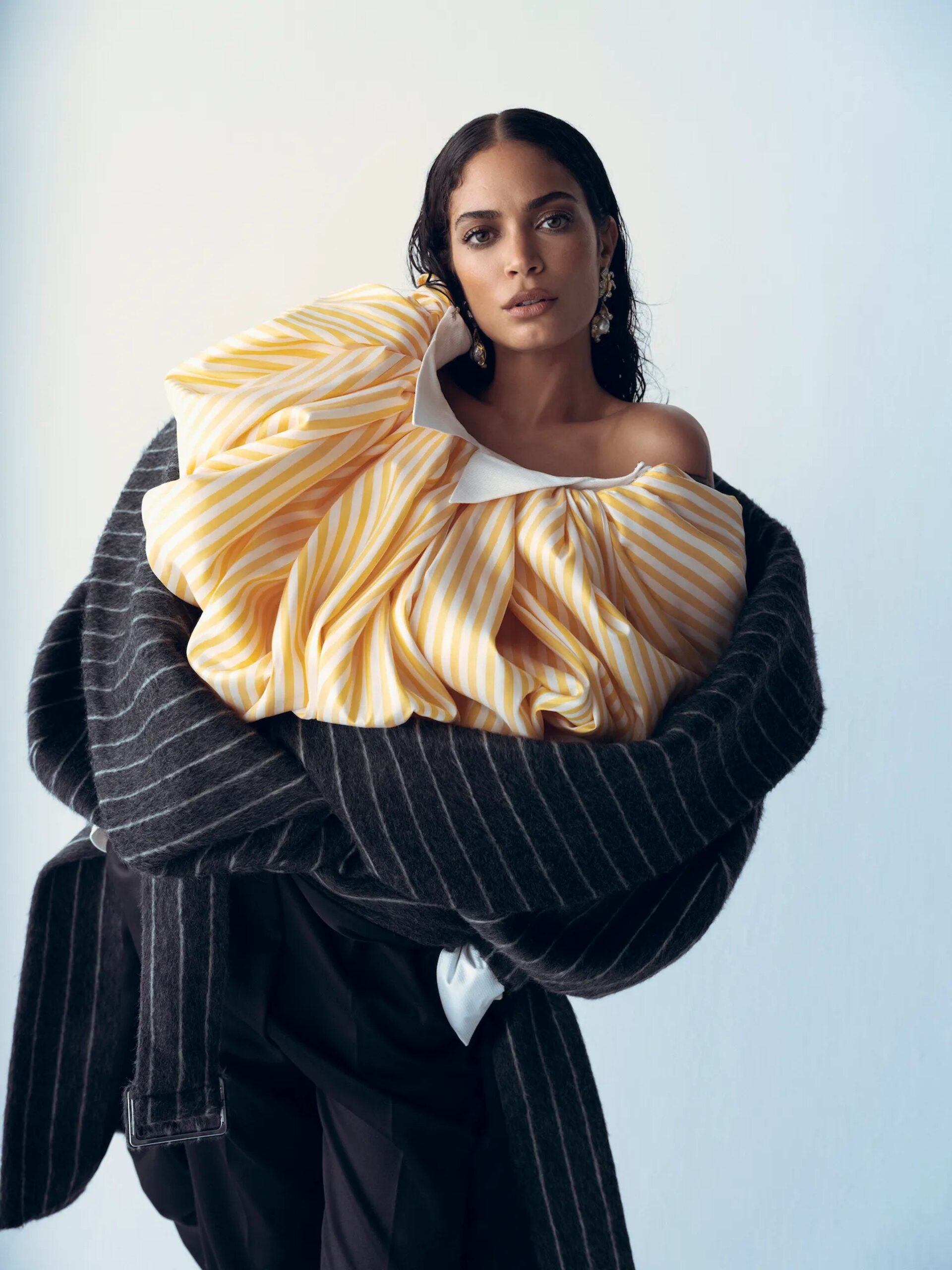 Model with dark wet-look hair wearing a voluminous yellow striped top and wrapped in a dark pinstripe coat.