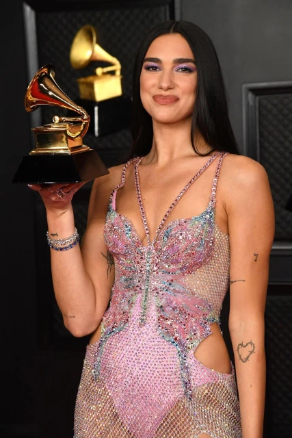 Dua Lipa holds a Grammy Award, wearing a pink and silver sparkly dress and dark eye makeup in a photo studio with a gray backdrop.