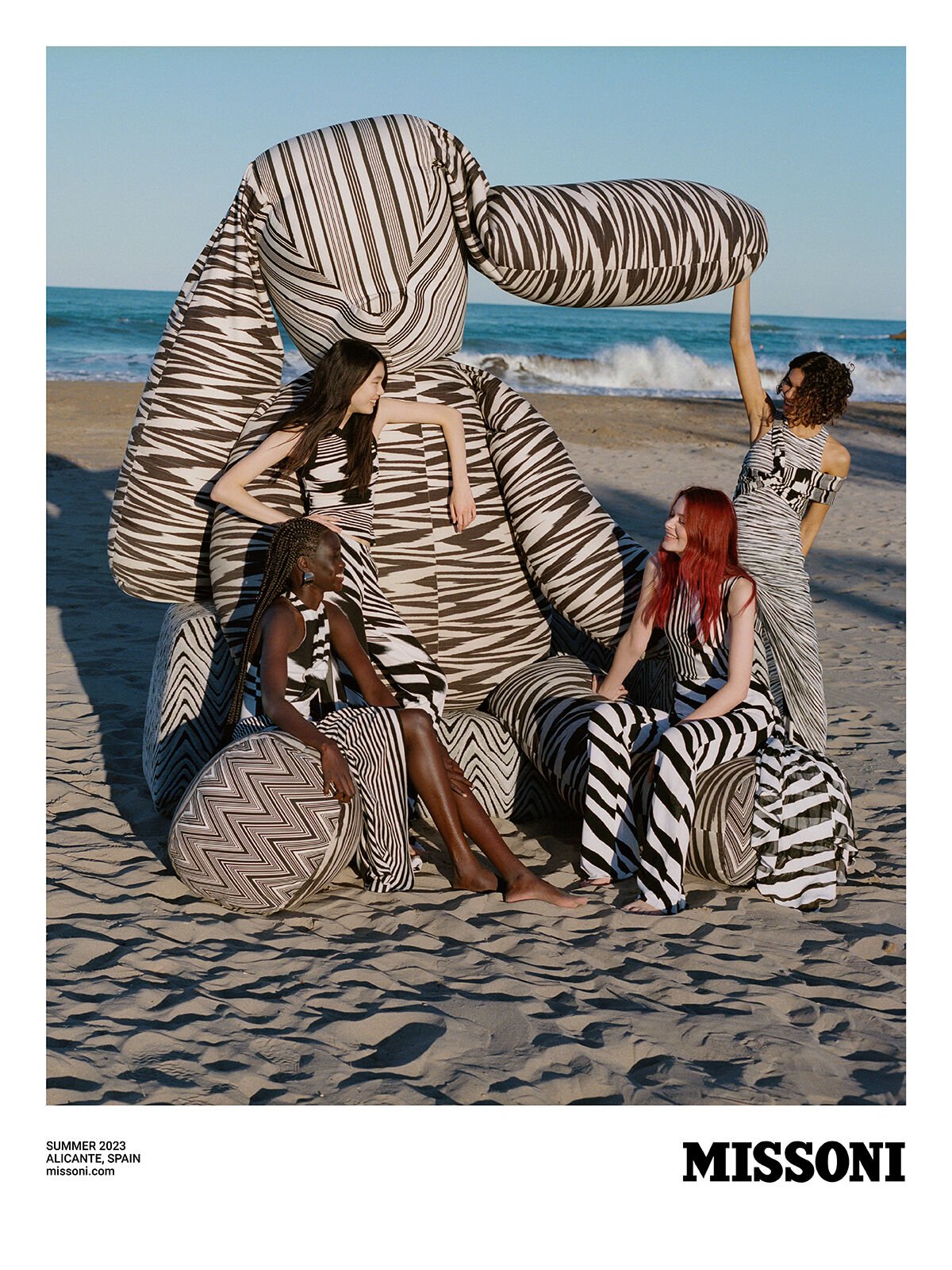 A group of four fashion models pose on a sandy beach beside a massive stuffed toy in an outdoor fashion editorial shot.