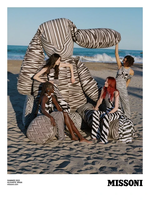A group of four fashion models pose on a sandy beach beside a massive stuffed toy in an outdoor fashion editorial shot.