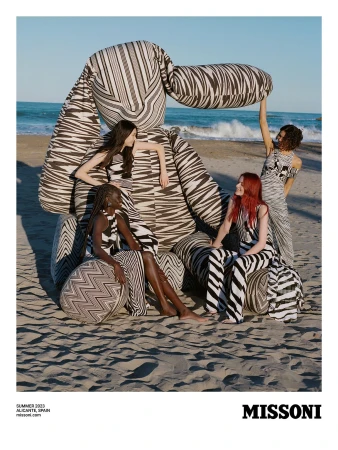 A group of four fashion models pose on a sandy beach beside a massive stuffed toy in an outdoor fashion editorial shot.