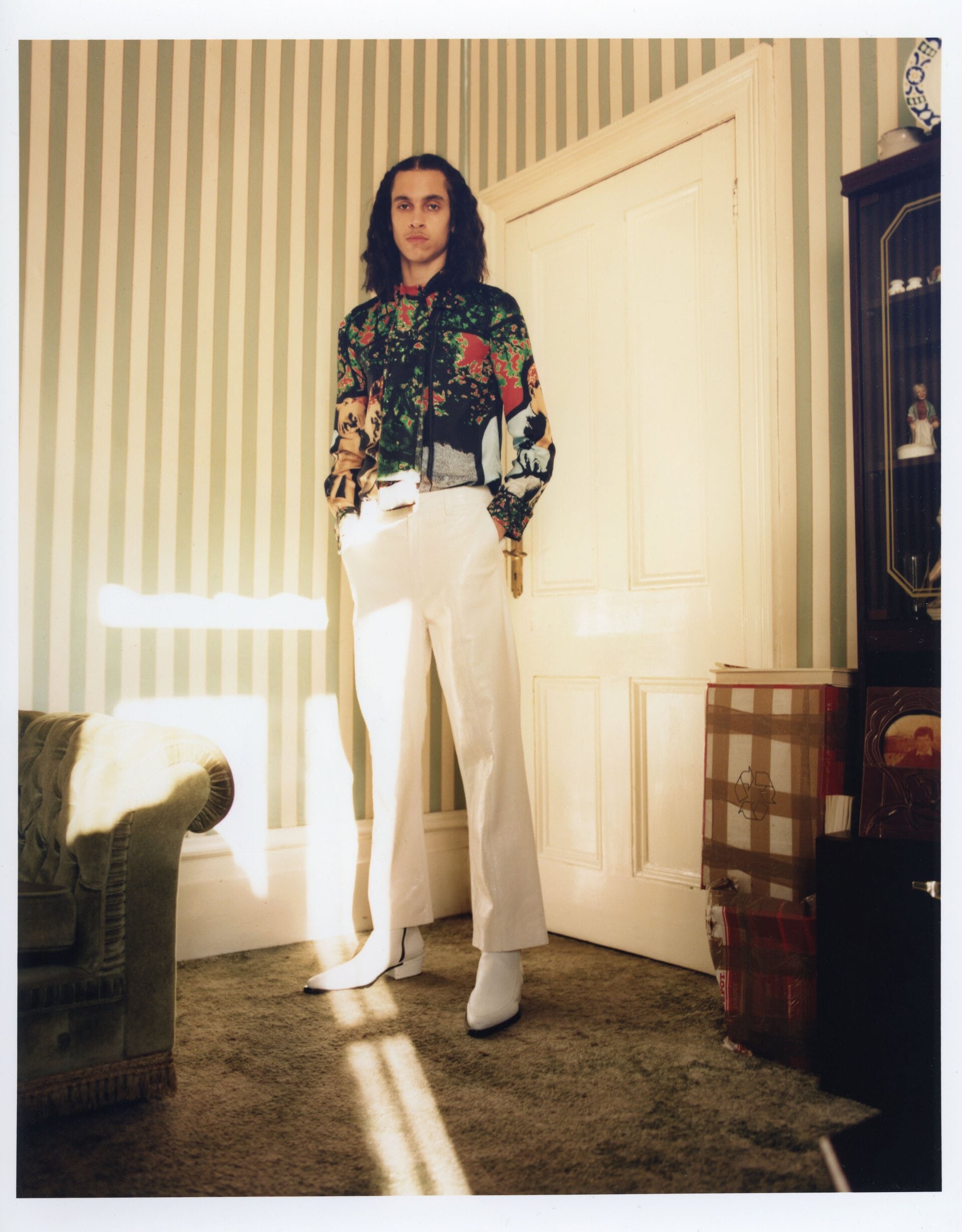 Adult male in a patterned shirt and white pants stands in a room with striped wallpaper and a vintage-style chair near a doorway.