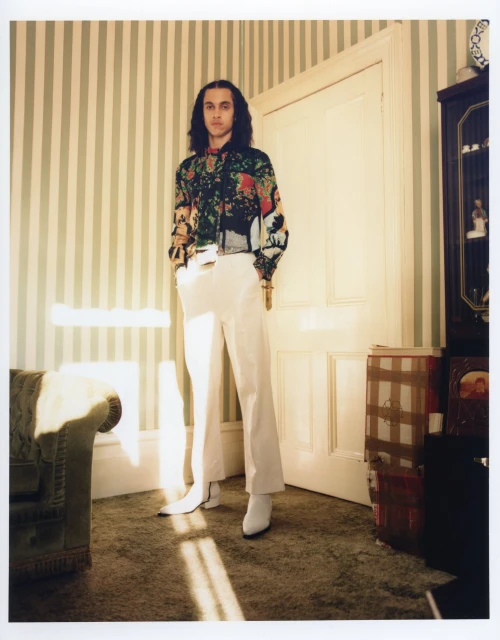Adult male in a patterned shirt and white pants stands in a room with striped wallpaper and a vintage-style chair near a doorway.