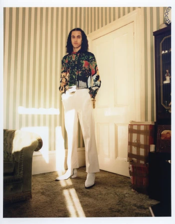 Adult male in a patterned shirt and white pants stands in a room with striped wallpaper and a vintage-style chair near a doorway.