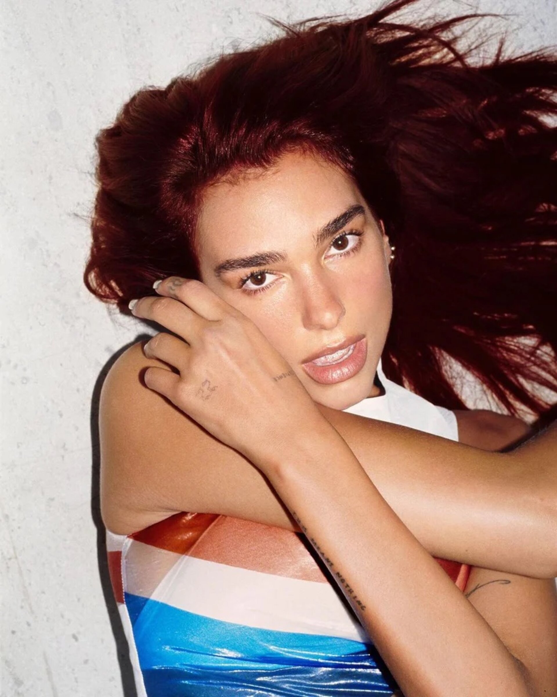 Headshot of a woman with maroon hair lying down, resting her head on her crossed arms and glancing at the camera against a grey background.