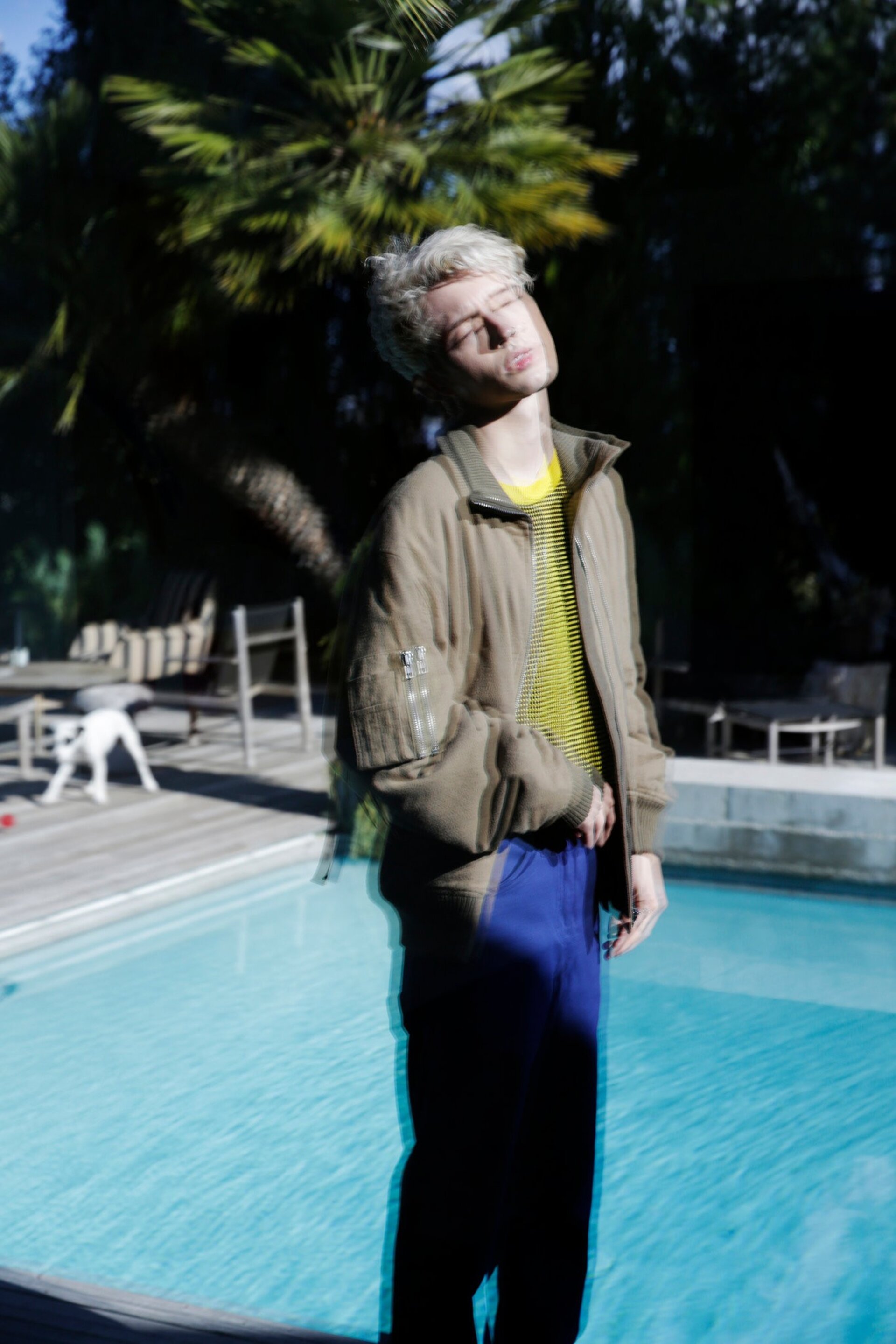 Young man poses near a pool in stylish khaki jacket, blue pants, and yellow striped shirt, bathed in natural sunlight with palm trees.