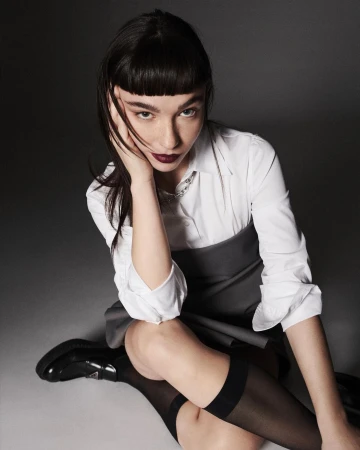 A seated woman wearing a white button-down shirt and grey dress styled by Lorenzo Posocco against a neutral grey studio background
