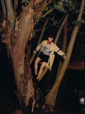 Young man in a beige patterned coat and shorts perched among large eucalyptus trees at night