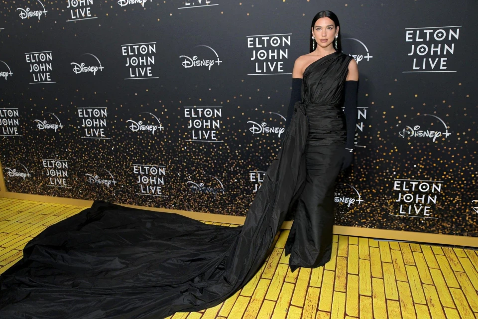 Dua Lipa poses in a black one-shoulder gown with a long train at the Elton John Live event on a yellow and black branded backdrop.