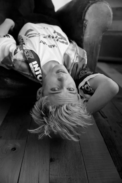 Monochrome portrait of a young adult with blonde hair lying upside down, wearing patterned t-shirt, in an indoor setting with wooden floor.