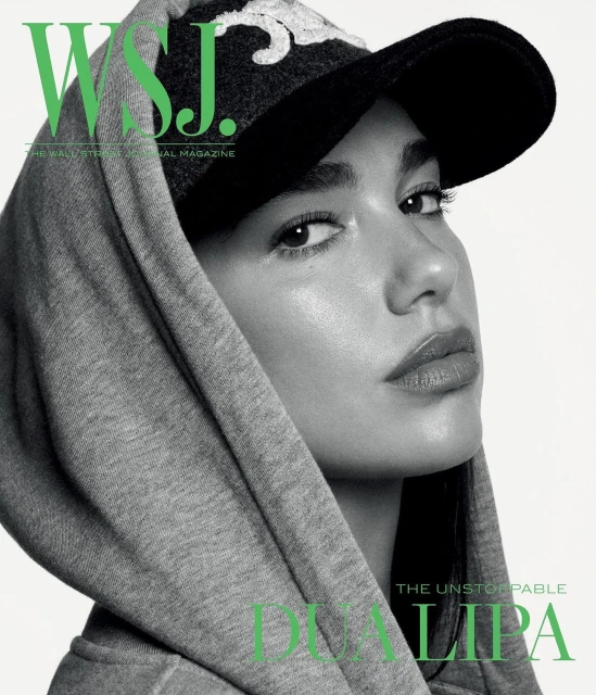 Close-up of Dua Lipa on a magazine cover, wearing a baseball cap and grey hooded sweatshirt against a bright white background.