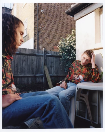 Two young adult males are seated in an outdoor setting near a fence, a building, and greenery, with one resting his head on his hand.