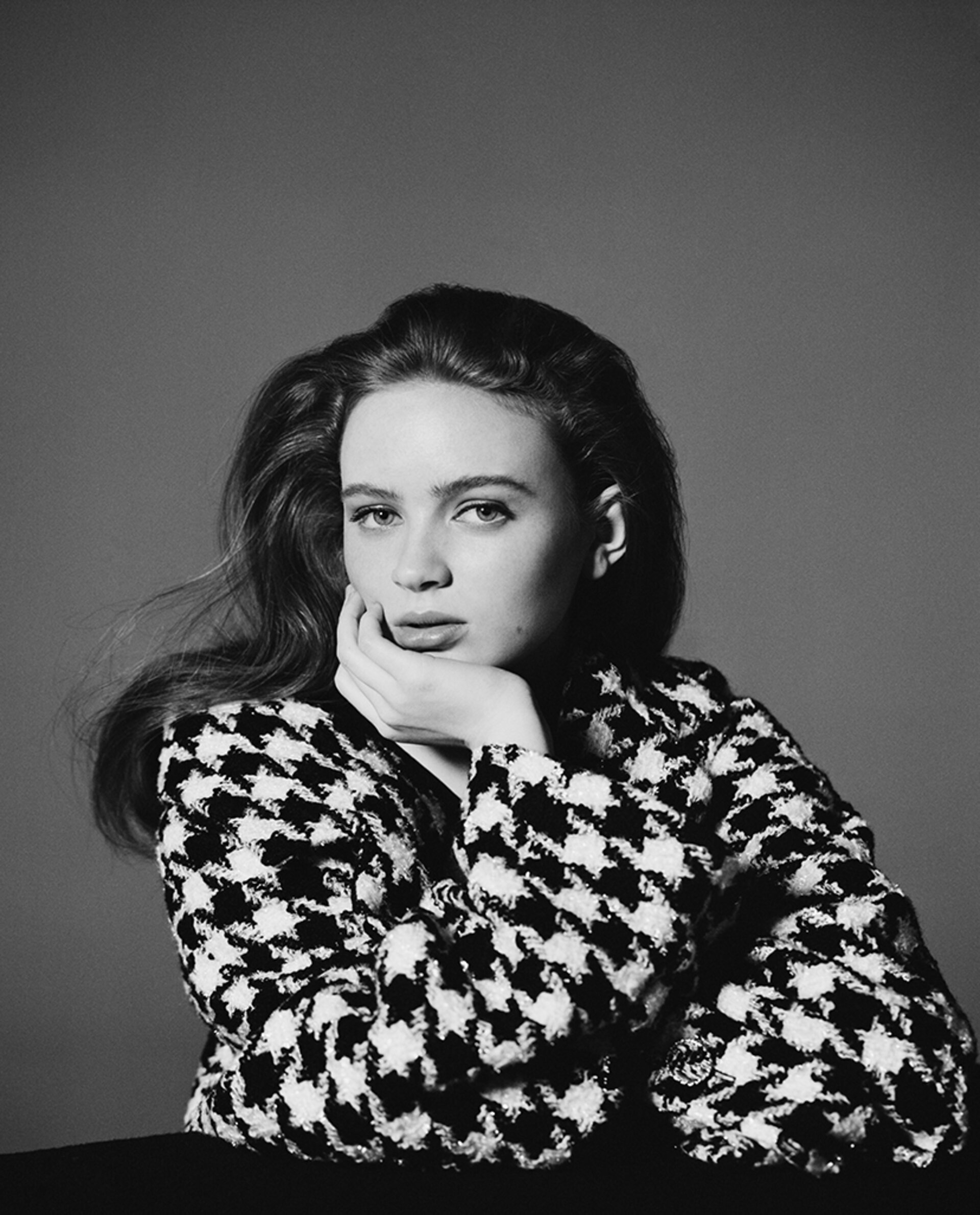 Monochrome portrait of a young adult female with long hair, wearing a houndstooth jacket, posing indoors against a grey backdrop.