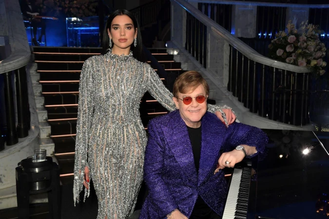 Dua Lipa and Elton John pose on a marble staircase with a piano, flowers, and stage lighting, showcasing a musical collaboration.