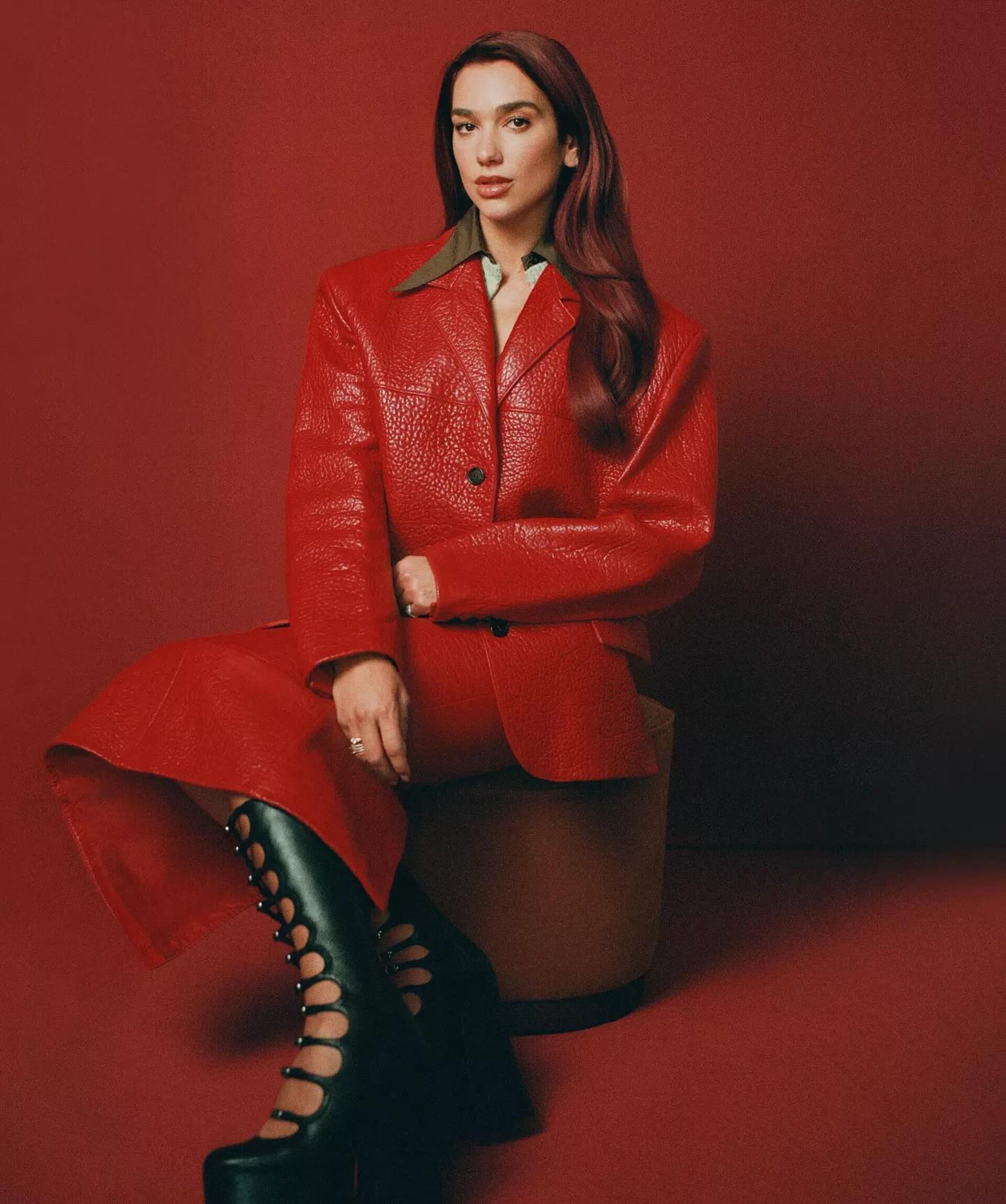 A stylish adult woman with red hair poses indoors wearing a red leather blazer, red skirt, and high-platform black boots against a red backdrop.