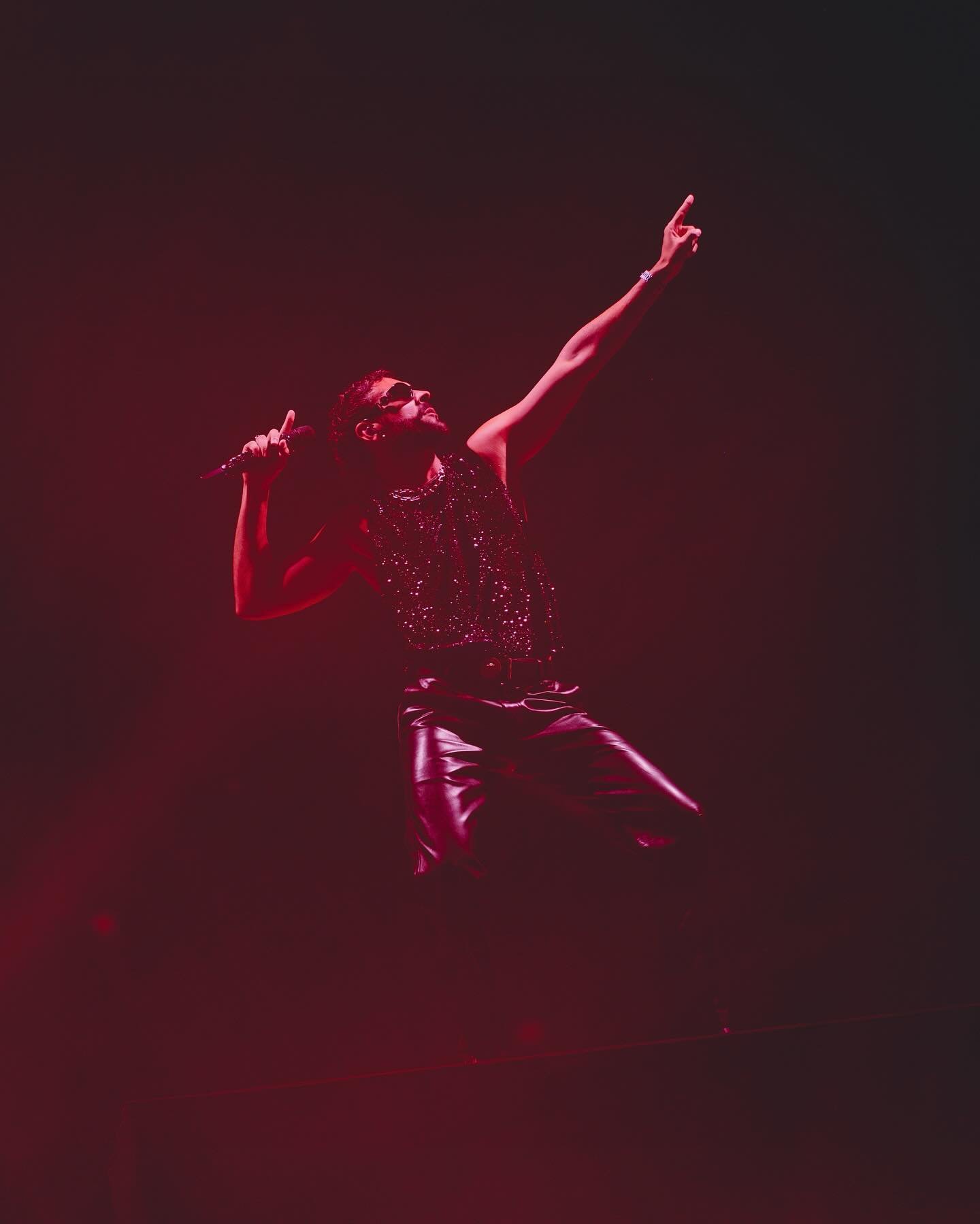 Male music artist sings on stage into a microphone during a live performance, bathed in red lighting.