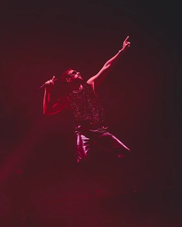 Male music artist sings on stage into a microphone during a live performance, bathed in red lighting.