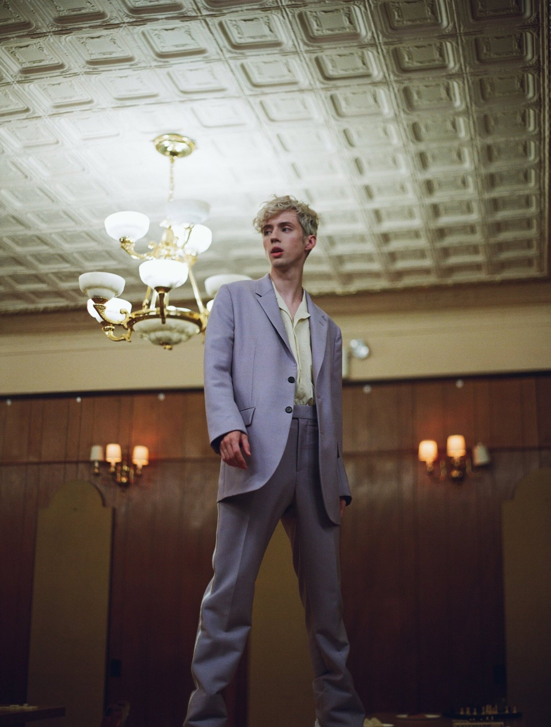 Young adult in a lilac suit stands beneath a chandelier. Indoor portrait showcases neutral tones and classic interior architecture.