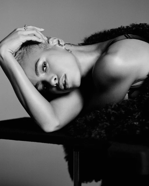 Portrait of a woman with short hair lying on a black surface, wearing a fur coat and jewelry in a black and white studio photograph.