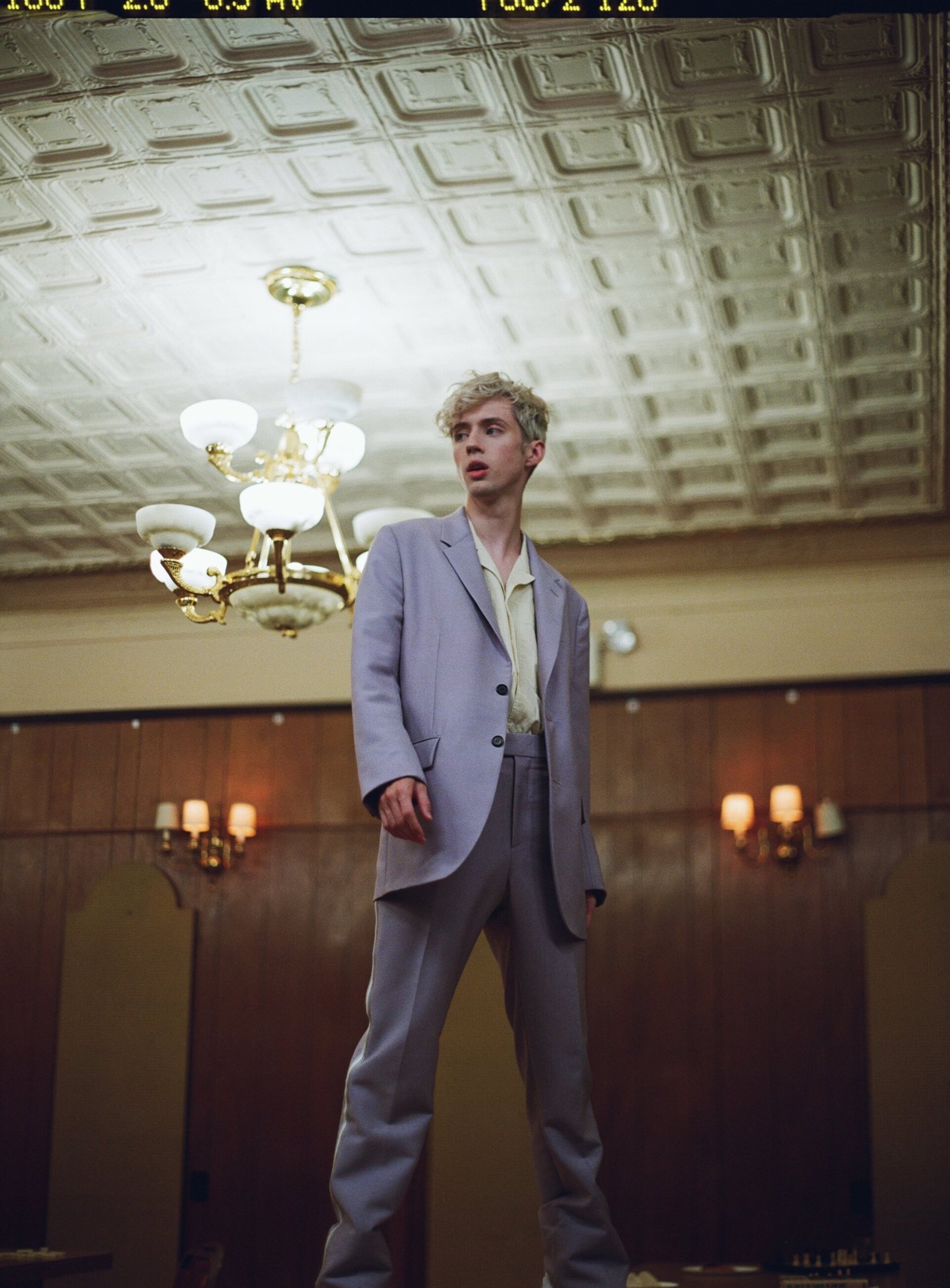 Young adult male in light purple suit stands under ornate chandelier in vintage ballroom interior with coffered ceiling and dark wood walls.