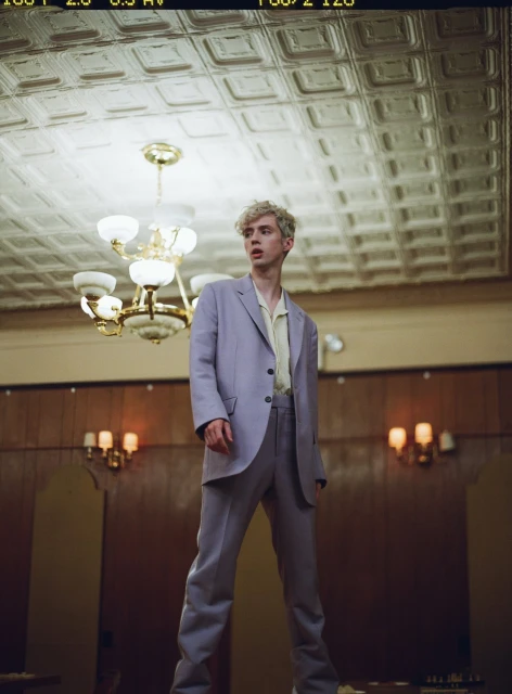 Young adult male in light purple suit stands under ornate chandelier in vintage ballroom interior with coffered ceiling and dark wood walls.