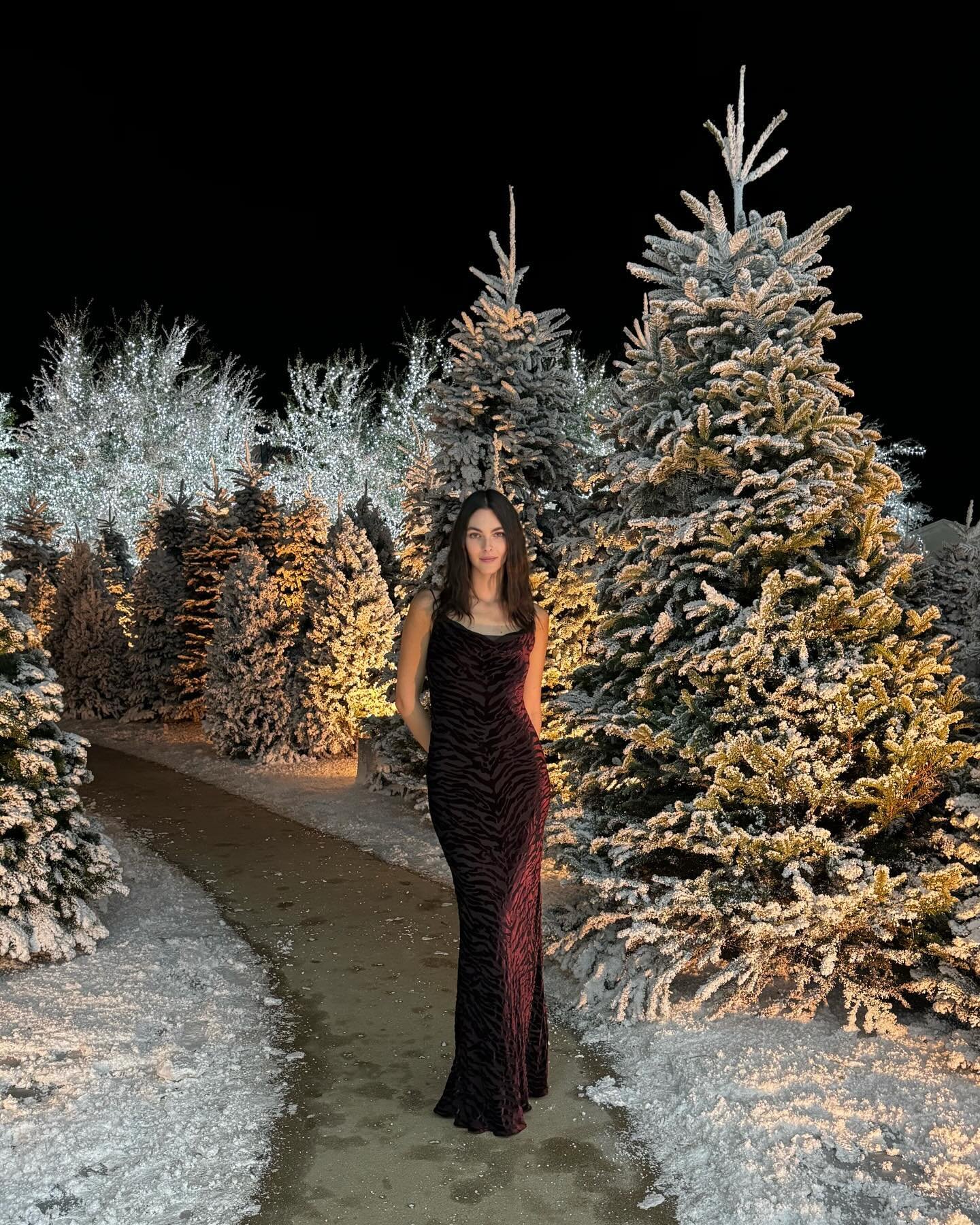 Elegant woman poses in a dark animal-print gown among snow-covered pine trees, creating a festive winter fashion scene at night.