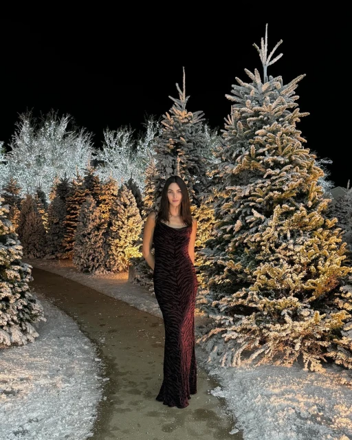 Elegant woman poses in a dark animal-print gown among snow-covered pine trees, creating a festive winter fashion scene at night.