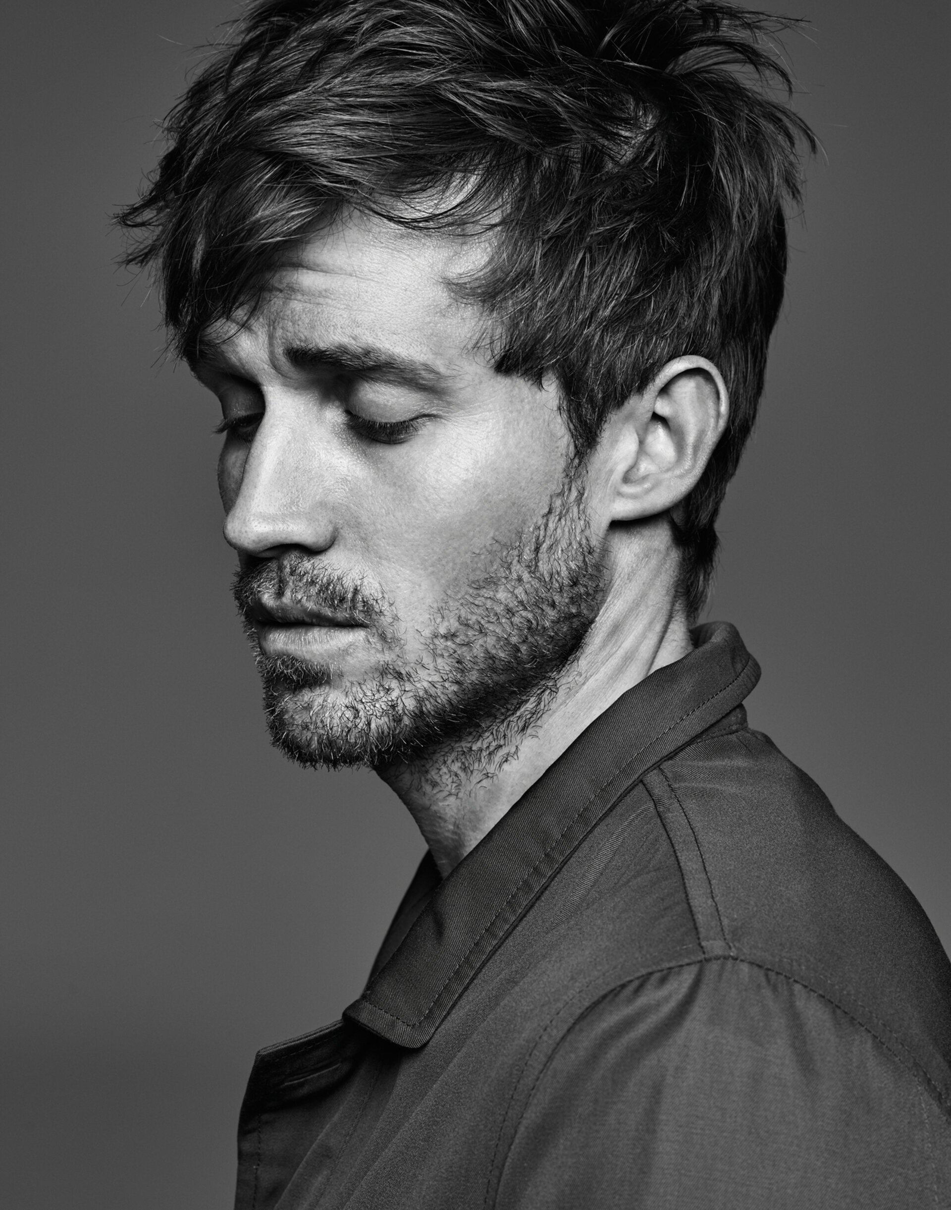 Monochrome portrait of a handsome adult male with dark hair, wearing a collared shirt, gazing downward with a contemplative expression in studio.