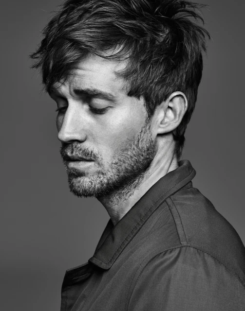 Monochrome portrait of a handsome adult male with dark hair, wearing a collared shirt, gazing downward with a contemplative expression in studio.