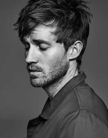 Monochrome portrait of a handsome adult male with dark hair, wearing a collared shirt, gazing downward with a contemplative expression in studio.