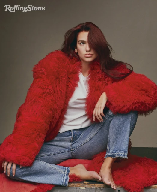 Portrait of Dua Lipa in a red faux fur coat and jeans, sitting barefoot against a grey backdrop for Rolling Stone magazine.