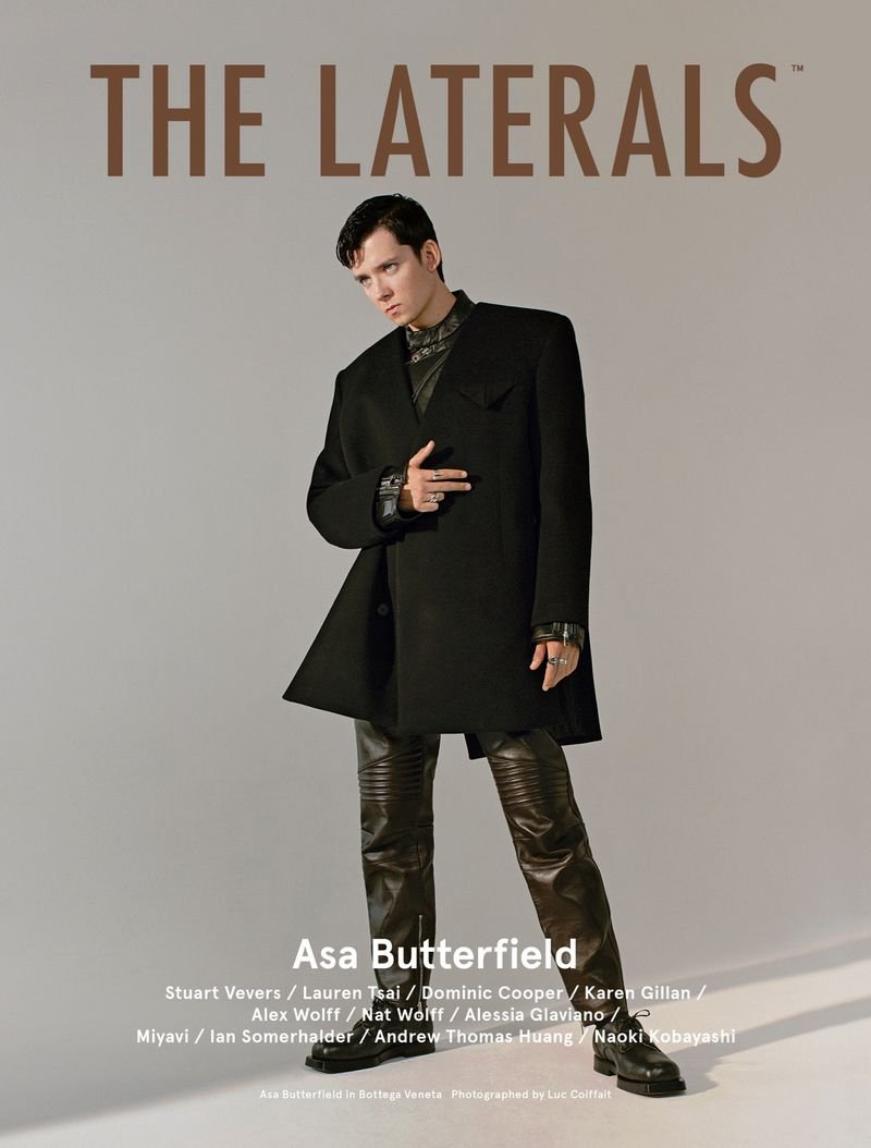 Asa Butterfield poses in a black coat and leather pants against a grey backdrop, featured on the cover of The Laterals magazine.